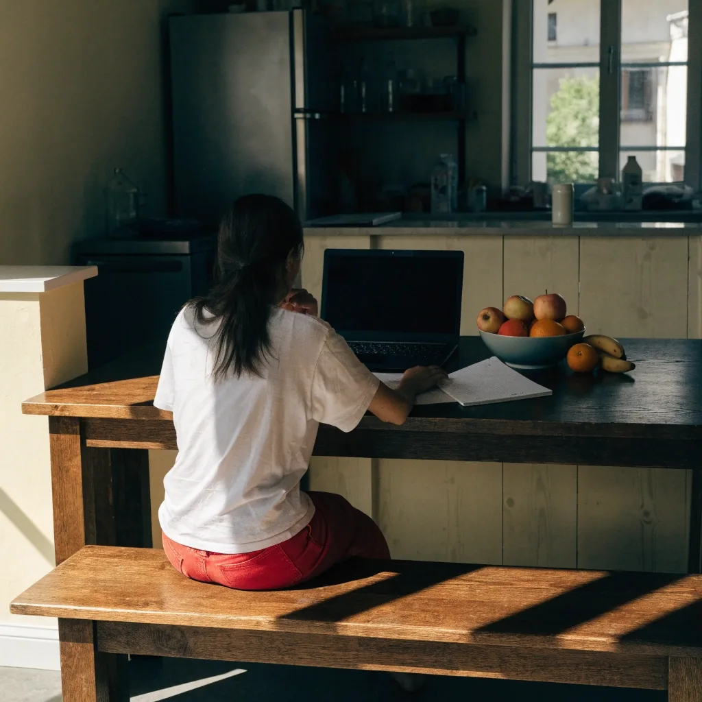 A woman at a kitchen bench in a video consultation, laptop angled away, a hand resting on a notepad beside it